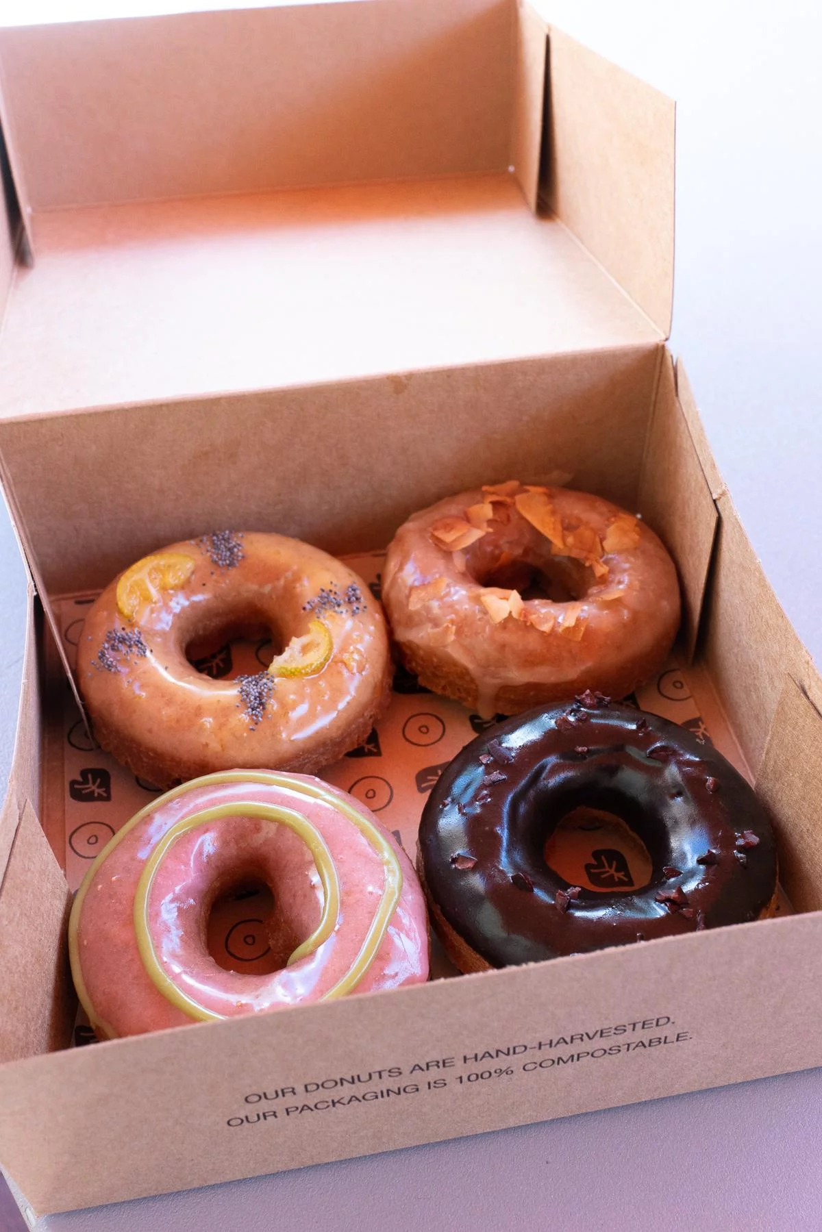 Opened box with four donuts from Holey Grail Donuts. The donut flavors are strawberry-green tea, Kauai chocolate and cacao nibs, maple and smoked coconut, and poppyseed and kumquats.