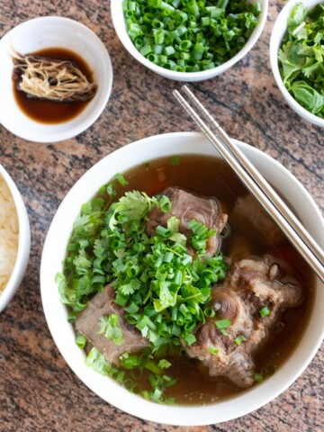 A bowl of oxtail soup with a side of rice, ginger and soy sauce for dipping, and green onions and cilantro to sprinkle on top.