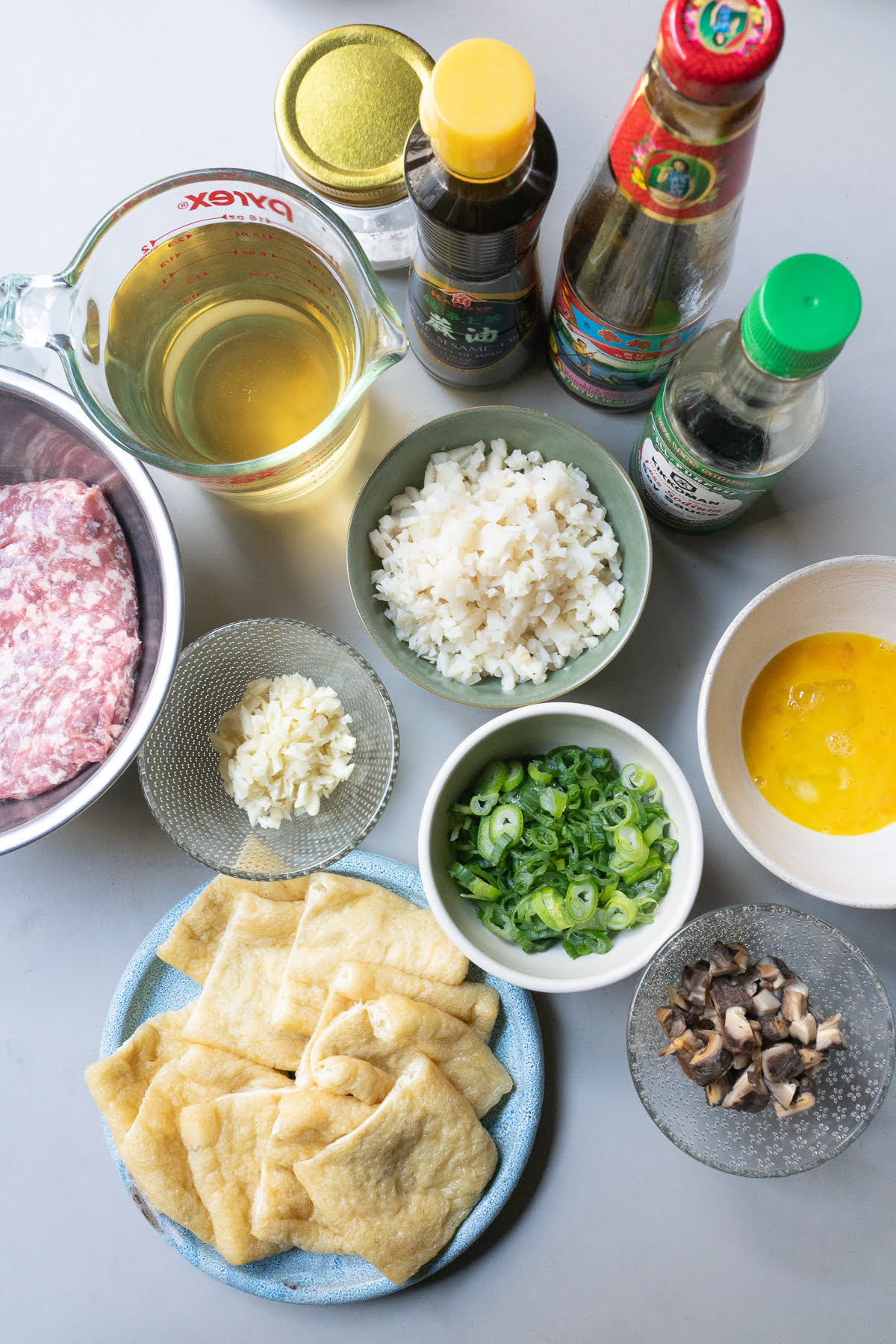Ingredients for stuffed aburage (aburage, ground pork, garlic, water chestnuts, shiitake mushrooms, green onions, egg, soy sauce, oyster sauce, sesame oil, cornstarch, and broth), laid out on a table.