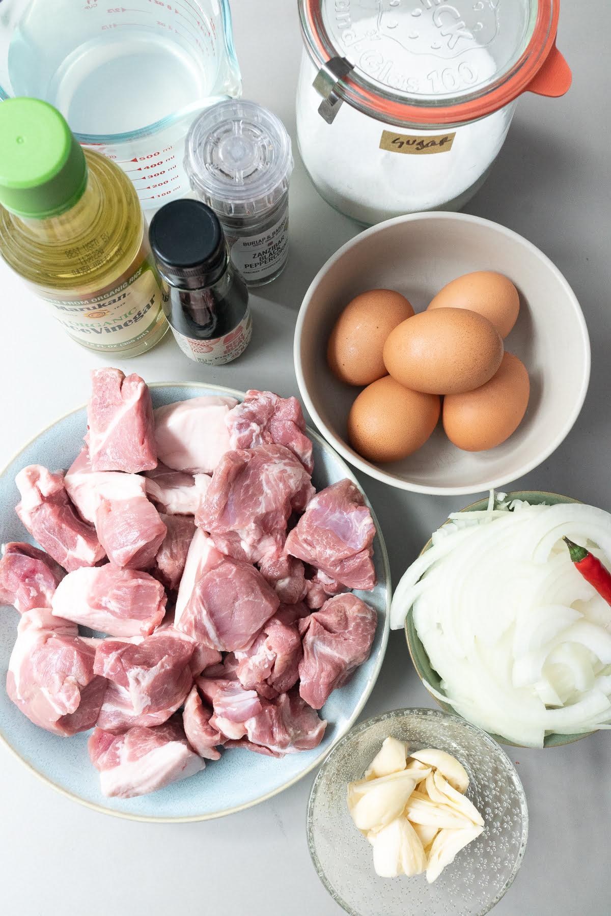 An overhead shot of the ingredients for Thit Kho (Vietnamese Braised Pork and Egg): pork shoulder, hard boiled eggs, garlic, onions, chili pepper, rice vinegar, coconut water, back peppercorns, fish sauce, and sugar.