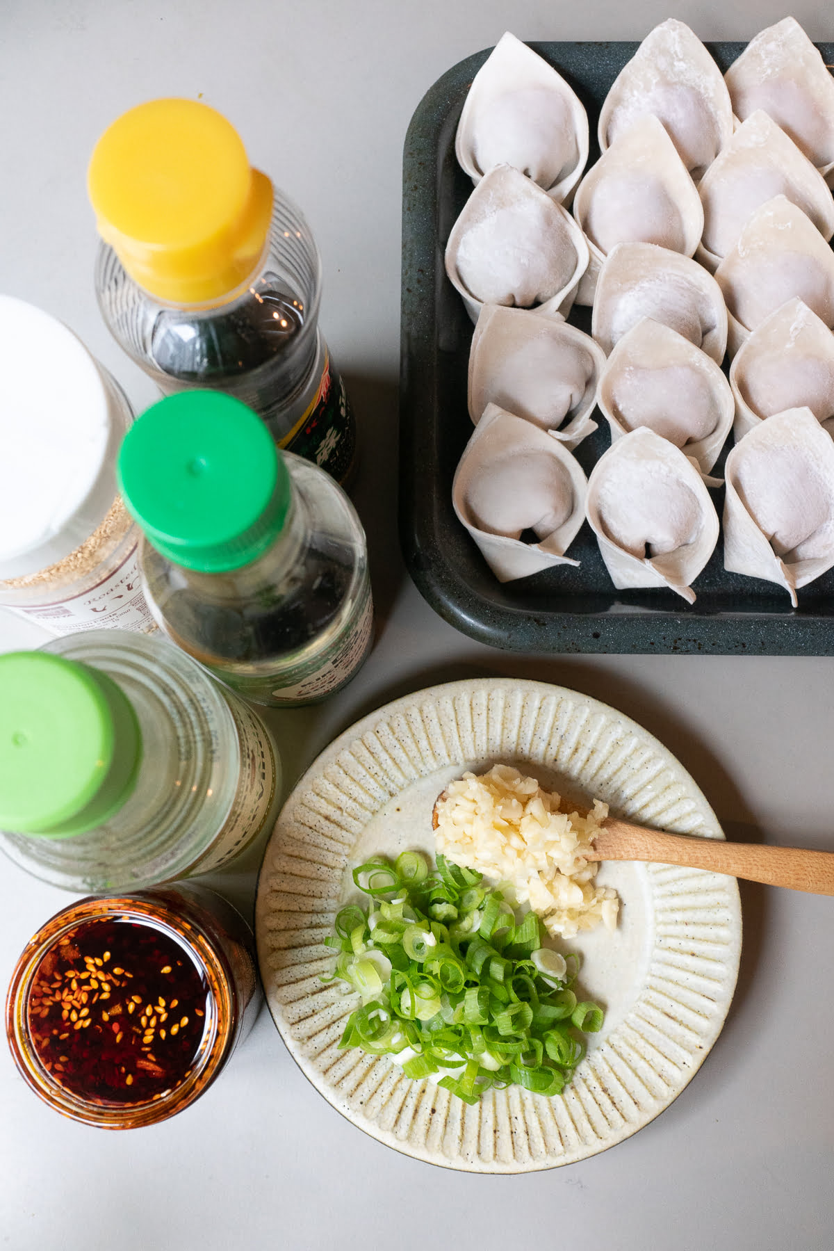 Ingredients for A plate of Szechuan Dumplings / Wontons on a table (wontons or dumplings, garlic, green onions, chili oil, soy sauce, rice vinegar, sesame oil, and sesame seeds).