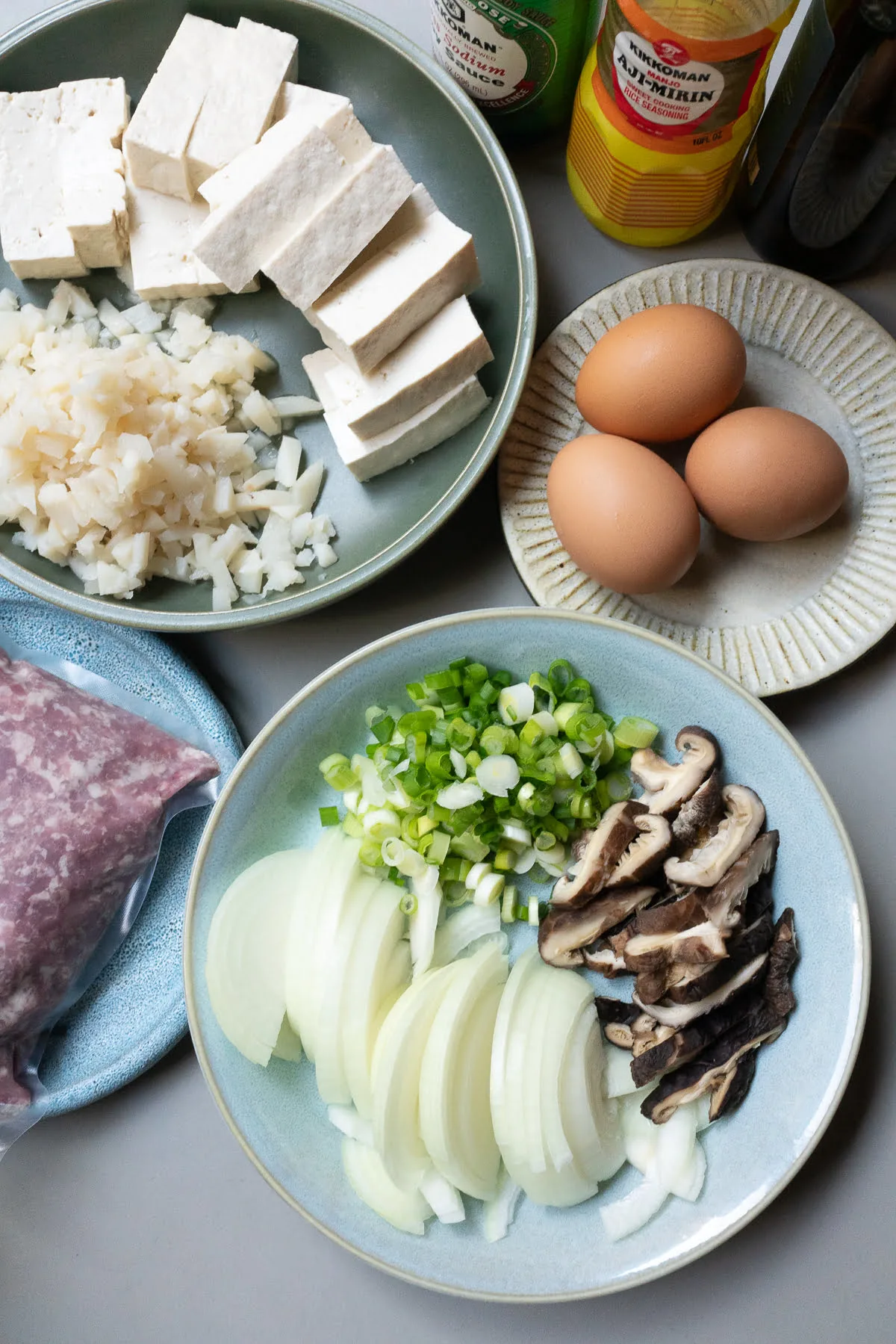 Ingredients for Pork Tofu Casserole (Taiwan Mushi) laid out on a table (ground pork, eggs, onions, mushrooms, green onions, tofu, water chestnuts, soy sauce, and mirin).