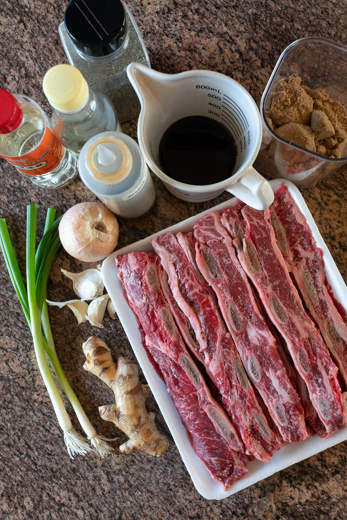 Ingredients for kalbi laid out on a table (soy sauce, water, sesame oil, brown sugar, mirin, rice vinegar, onion, garlic, ginger, and green onions).