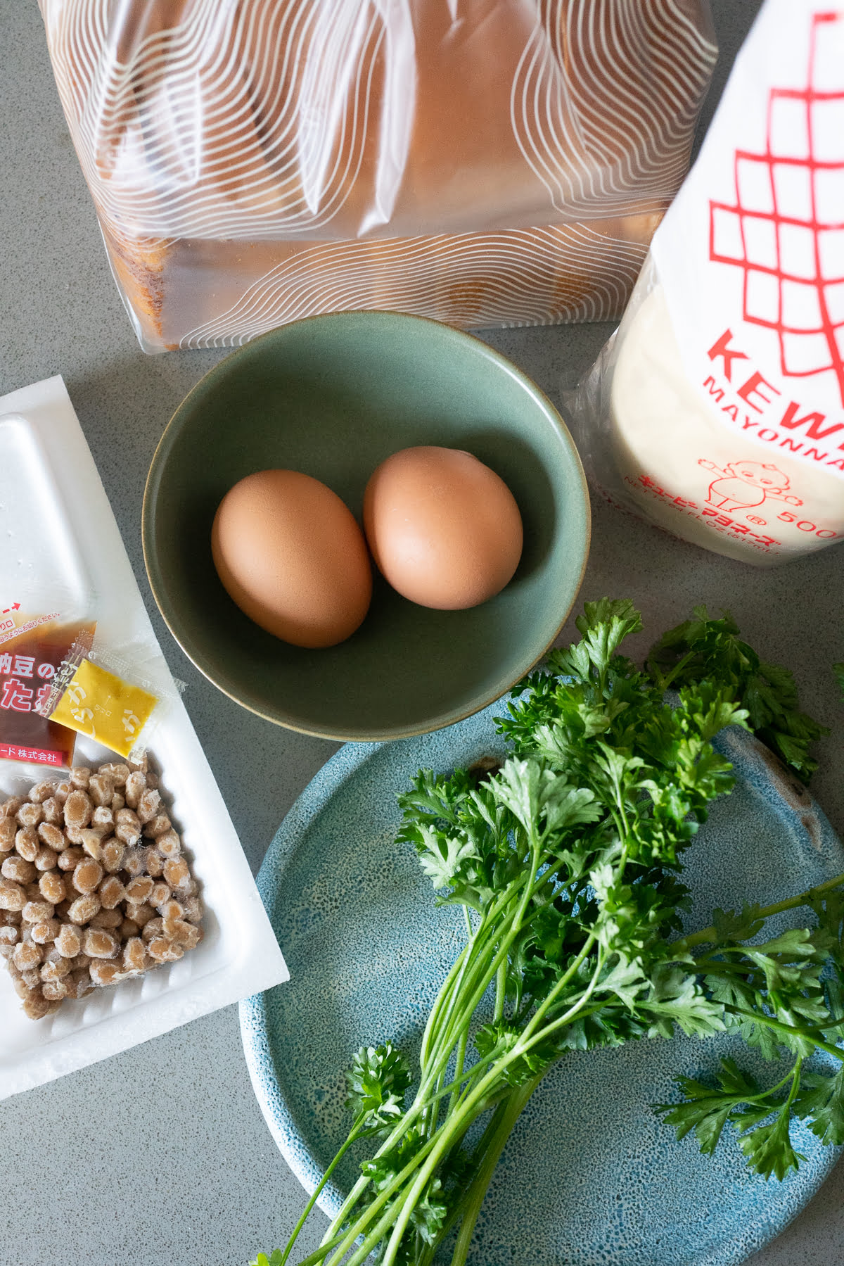 Ingredients for Natto Egg Toast on a counter (bread, mayo, eggs, parsley, and natto).