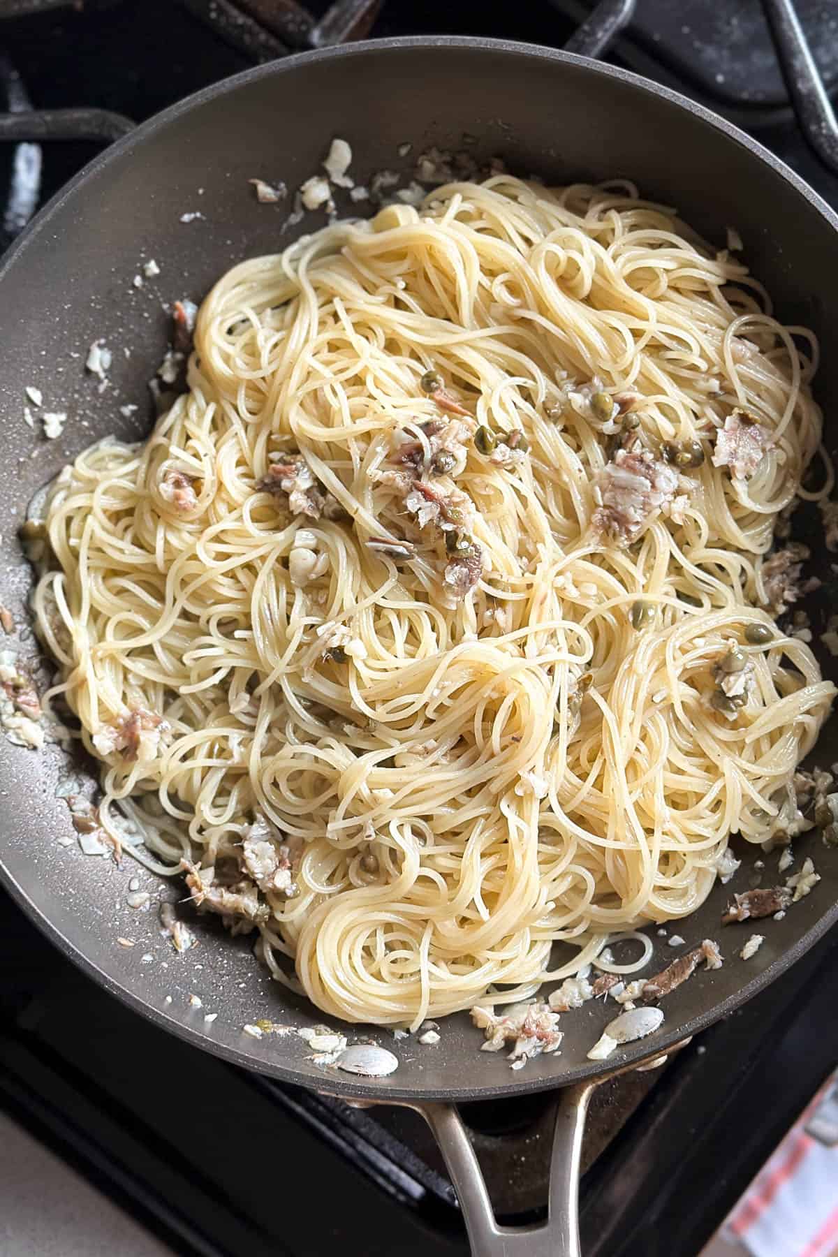 Mixing together angel hair pasta with anchovies, garlic, and capers in a pan.