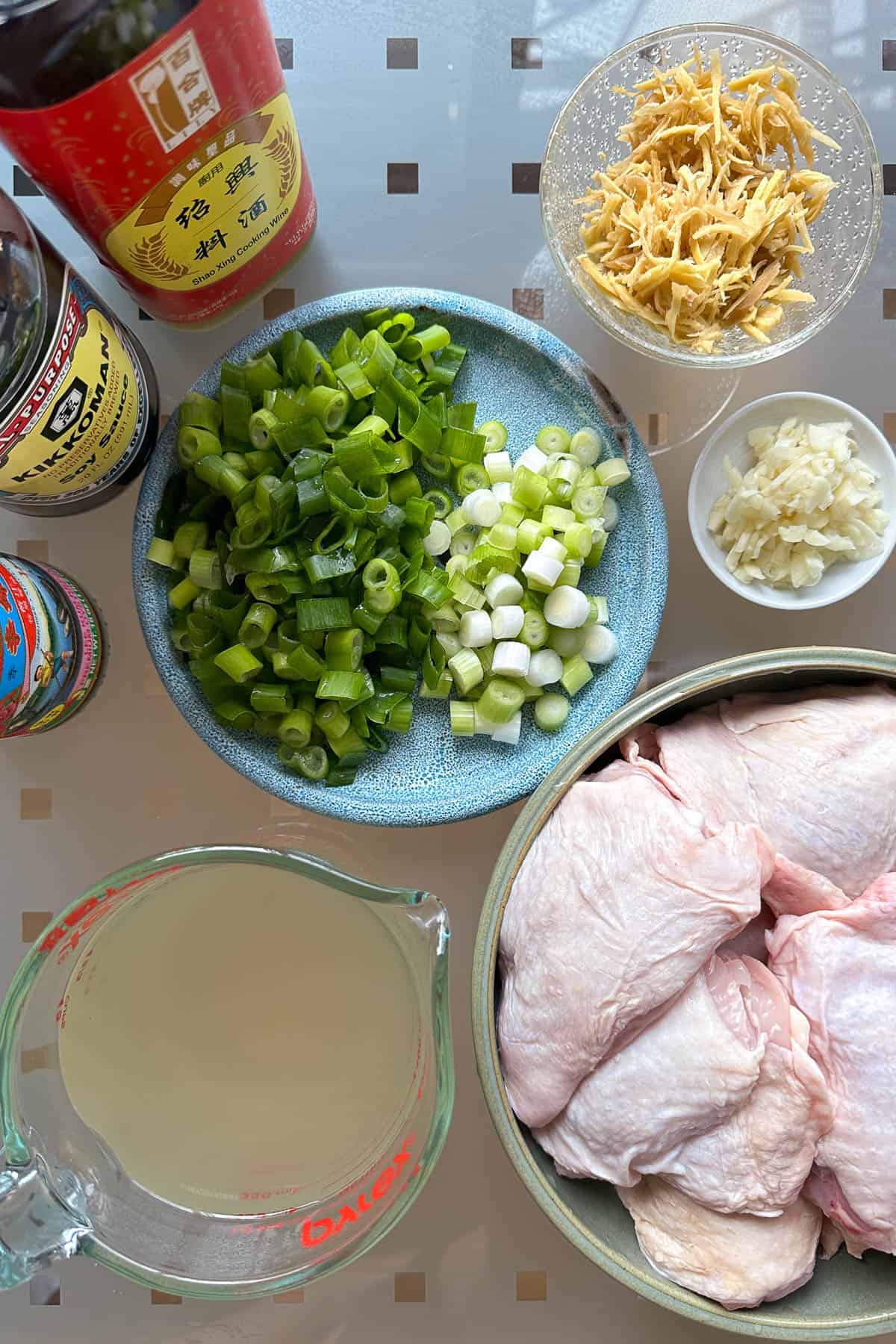 Ingredients for oyster sauce chicken laid out on a table (chicken thighs, chicken broth, green onion, ginger, garlic, oyster sauce, Shaoxing wine, and soy sauce).