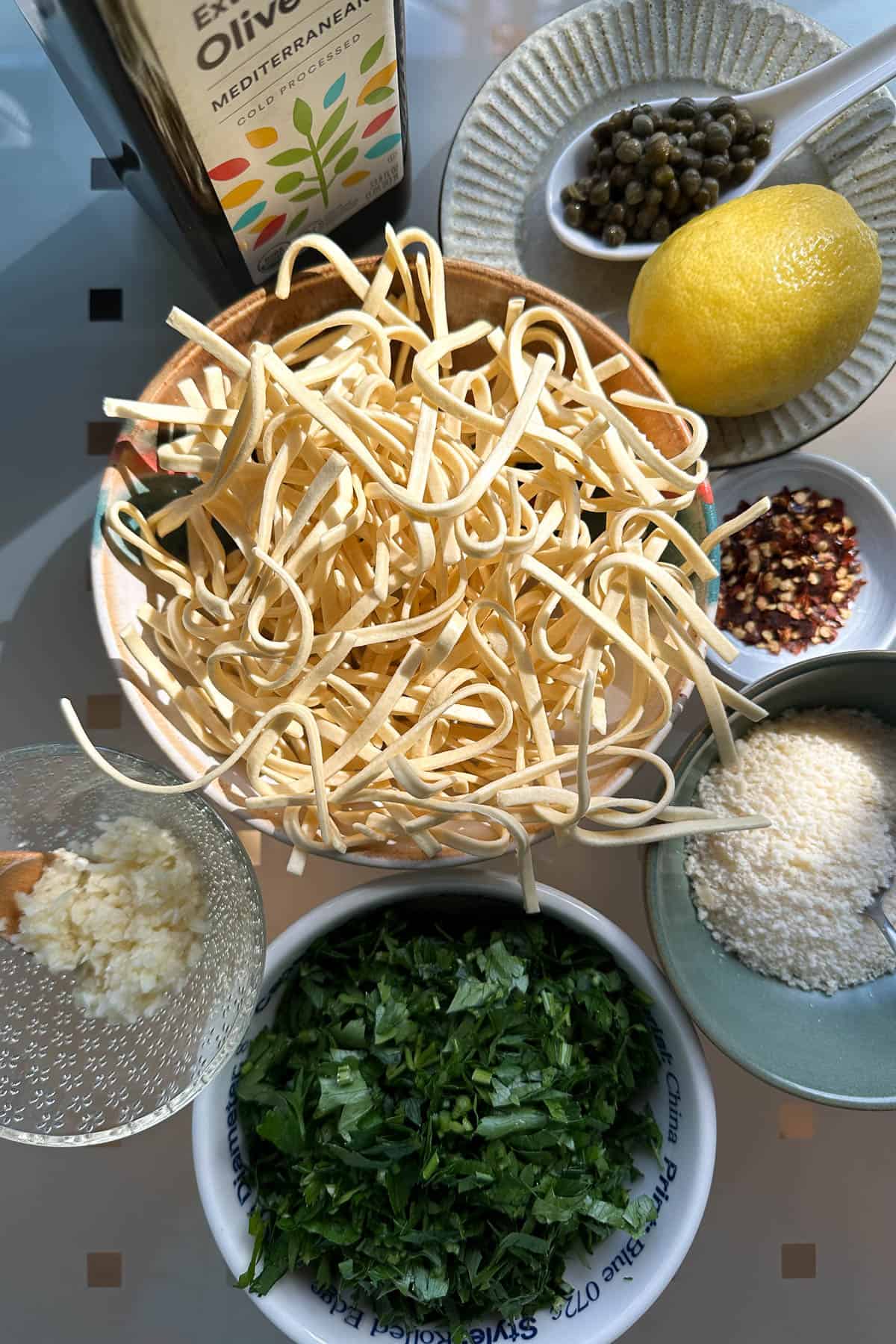 Ingredients for parsley pasta laid out on a table (parsley, pasta, olive oil, chili flakes, capers, garlic, lemon, and parmesan cheese).