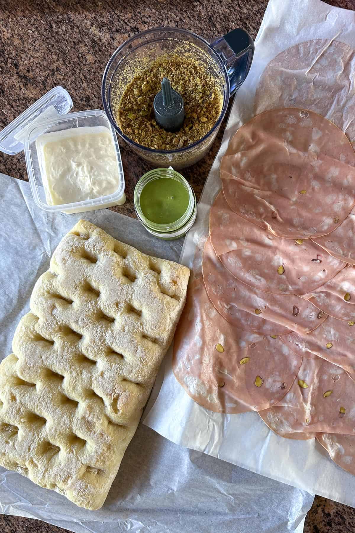 Ingredients for Mortadella Sandwich on a counter (focaccia, mortadella, pistachio paste, stracciatella, crushed pistachios).